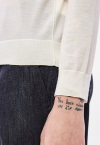 Pulover tricotat bej cu manșete striate, asortat cu blugi din denim închis. Tatuaj vizibil cu text pe încheietură. Prim-plan concentrat pe țesătură și detaliile designului.