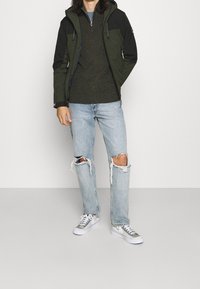 Grön och svart huva jacka, stickad olivgrön tröja, ljusblå slitna jeans och grå/silver sneakers med vita sulor.