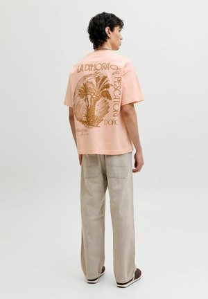 Hátrafelé néző férfi, aki barackszínű pólót visel barna pálmafa mintával és szöveggel, bézs nadrágot és sötét sportcipőt fehér háttéren.