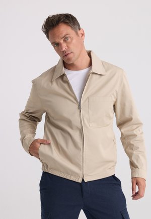 Homme portant une veste beige zippée sur une chemise blanche et un pantalon bleu marine, une main dans une poche sur un fond clair uni.