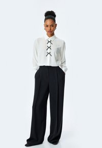 Blusa blanca con cuello, con cintas de raso negro en el frente, combinada con pantalones de pierna ancha negros de alta cintura.