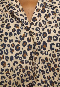 Chemise beige avec motif léopard noir et marron, boutons noirs et une poche poitrine.