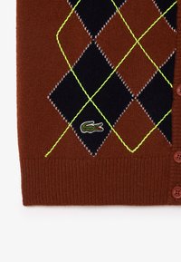 Cardigan lavorato a maglia marrone con un motivo argyle nero e marrone, cuciture in neon verde e un piccolo logo ricamato. Sei bottoni.