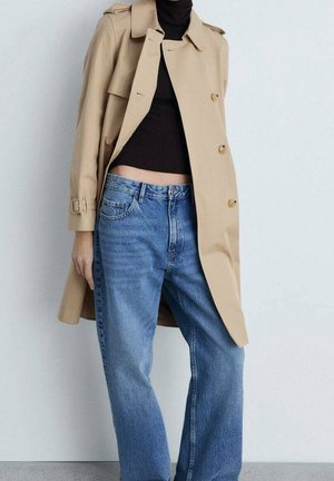 Personne portant un trench beige sur un col roulé court noir et un jean bleu taille haute, se tenant devant un fond blanc uni.