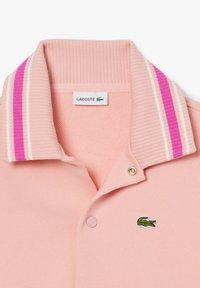 Světle růžové polo tričko s žebrovaným límcem, na kterém jsou růžové a bílé pruhy, se zlatým zapínáním na knoflík a vyšitým zeleným krokodýlem.