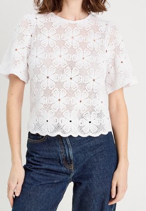 Kvinde iført en hvid kortærmet blondebluse med blomsterprint kombineret med mørkeblå højtaljede jeans.