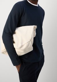 Granatowa koszula z długimi rękawami noszona przez osobę, trzymającą kremową, luźną torbę z płótna z zagiętym wierzchem i pętelką. Teksturowana tkanina i minimalistyczny design.