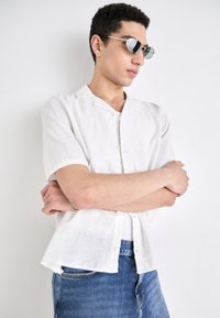 Fehér vászon, rövid ujjú gombos ing, gallérral. Kék farmernadrággal párosítva. A modellen tükrös napszemüveg van.