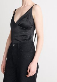Svart satin bodysuit med djup V-hals, justerbara spaghettistövlar och CK-logotyp; ihop med högmidjade svarta jeans med rhinestone-accenter.