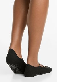 Black ballet flats made of soft fabric, featuring a curved shape, elastic straps, and a textured sole with grip pattern for stability.