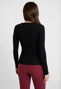 Donna con lunghi capelli castani che indossa un top nero a coste a maniche lunghe e jeans bordeaux, rivolta di spalle contro uno sfondo chiaro e semplice.