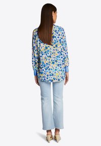 Bloes met bloemenprint in blauw, geel en wit op een lichte achtergrond, met lange mouwen en een ronde Zoom. Gepaard met lichte jeans.