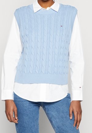 Personne portant un pull sans manches bleu clair en maille torsadée par-dessus une chemise blanche boutonnée et un jean bleu, debout les bras détendus.