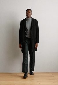 Manteau noir avec des boutons, col roulé gris et pantalon vert foncé. Porté avec des chaussures noires, sur un fond neutre.