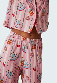 Rosa pyjamas set med en rolig tårta och körsbärstrykt. Mjuk tyg, kortärmad topp med knappar och byxor med elastisk midja.