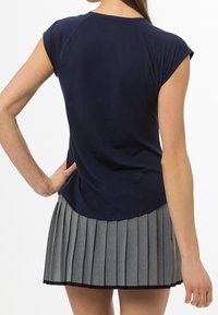 Kvinna sedd från baksidan, iklädd en marinblå t-shirt med kort ärm och en grå plisserad kjol, med en hand på höften.