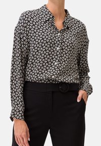 Camicia a bottoni nera con un motivo geometrico bianco; maniche lunghe con polsini a bottone; abbinata a pantaloni neri a vita alta e cintura.