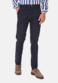 Pantaloni di cotone blu navy con vestibilità dritta, dotati di chiusura con bottone e tasche laterali, abbinati a scarpe in suede marrone.