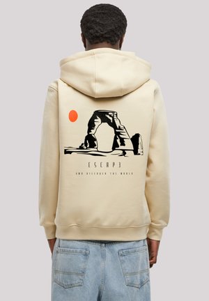 Beige Kapuzenpullover mit einem großen Grafiken eines Bogens und einer roten Sonne auf der Rückseite. Der Text lautet "FLIEH UND ENTDECKE DIE WELT" in Schwarz. Weicher Stoff.