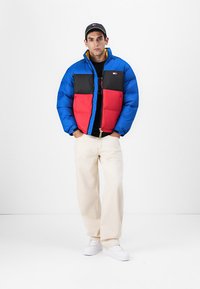 Gevoerde jas met blauwe, rode en zwarte panelen, voorzien van een rits en zakken. Draag je over een zwart t-shirt en gecombineerd met crèmekleurige broek en witte schoenen.