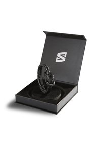 Black braided leather bracelet displayed in a black box with a soft, circular cushion. The box features a logo on the lid.