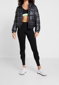 Černá péřová bunda s opakujícím se bílým logem, spárovaná s černým sportovním podprsenkou s zeleným pruhem a černými legínami se zelenými akcenty.