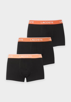 Jeune homme debout pieds nus portant un boxer noir avec une ceinture orange portant la mention Lacoste sur un fond clair uni.