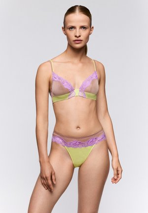 Frau trägt ein transparentes Lavendelfarbenes Spitzen- und hellgrünes Dessous-Set, steht vor einem einfachen hellgrauen Hintergrund und schaut geradeaus.