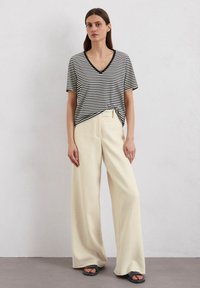 Gestreiftes schwarz-weißes V-Ausschnitt-T-Shirt, am Bund geknotet, kombiniert mit cremefarbenen weit geschnittenen Hosen und schwarzen Sandalen. Glatte Texturen überall.