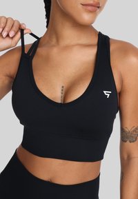 Zwarte sportbeha van rekbaar materiaal met een diepe V-hals, verstelbare bandjes en een wit logo aan de linkerkant.