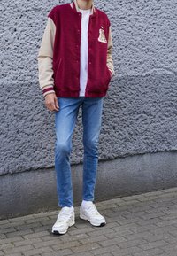 Vinröd och beige fleeceskjorta med bokstavsfläck, kombinerad med ljusblå jeans och vita sneakers med gröna detaljer.