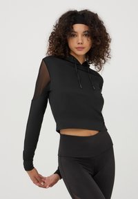 Cropped hoodie nera con maniche lunghe, caratterizzata da dettagli in rete sulle spalle e un cappuccio con cordino. Abbinata a leggings neri a vita alta.