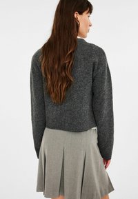 Eine Frau mit langen braunen Haaren, die einen dunkelgrauen Pullover und einen hellgrauen gefalteten Rock trägt, sieht von hinten gegen einen schlichten Hintergrund.