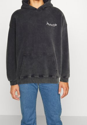 Osoba ubrana w ciemnoszarą bluzę z kapturem z przednią kieszenią oraz napisem "Muhammad Ali THE GREATEST" na piersi, w zestawie z niebieskimi dżinsami.