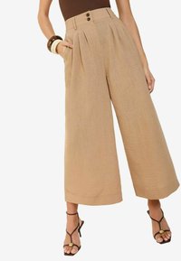 Beige weit geschnittene Hosen mit Falten, zwei Knopfverschluss an der Taille und Taschen. Der Stoff hat eine strukturierte Oberfläche, und der Saum ist cropped.