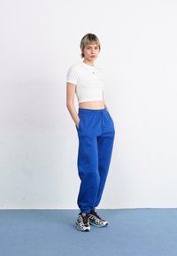 Blaue Sporthose mit elastischem Bund, kombiniert mit einem weißen cropped T-Shirt und schwarz-weißen Sneakern mit Muster. Einfaches und lässiges Design.