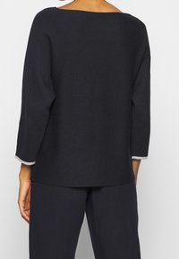 Czarny dzianinowy top z szerokim dekoltem w kształcie łódki, rękawami o długości do łokcia i kontrastowym jasnoszarym obszyciem przy mankietach. Gładka faktura, luźny krój.