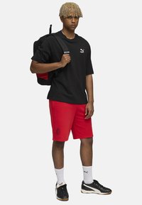 Camiseta negra de gran tamaño, pantalones cortos deportivos rojos con logo, calcetines blancos y zapatos negros con detalles blancos. Mochila con detalles rojos.