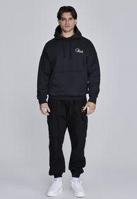 Sudadera negra con bolsillo frontal y capucha con cordón, combinada con pantalones cargo negros y zapatillas blancas. Textura de tela suave.