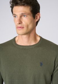 Maglione verde lavorato a maglia con scollo rotondo e un piccolo logo blu navy sul lato sinistro del petto. Texture liscia e cuciture uniformi in tutto il capo.