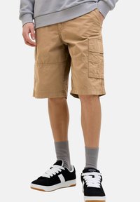 Beige Cargo-Shorts aus Baumwolle, mit Seitentaschen, einem lässigen Schnitt und umgeschlagenen Säumen, kombiniert mit schwarzen Sneakers und grauen Kniestrümpfen.