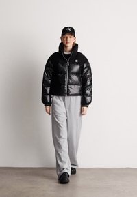 Melnā pūkainā jaka ar spīdīgu tekstūru un rāvējslēdzēju, apvienota ar pelēkiem treniņbiksēm, melnu cepuri un melnām slīdošajām kurpēm.