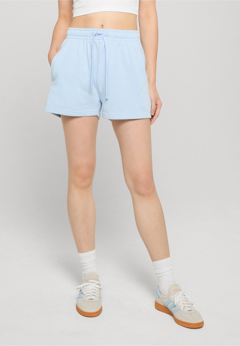 Femme portant un short bleu clair avec cordon, un crop top blanc, des chaussettes blanches et des baskets beige avec des rayures bleues, debout avec une main dans la poche.