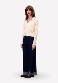 Vrouw met golvend bruin haar, gekleed in een crèmekleurige trui met lange mouwen en kraag, een brede donkerblauwe jeans met omslagen en bruine loafers, staand tegen een witte achtergrond.