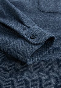 Manșeta cămășii cu model herringbone de culoare bleu închis, cu o textură fină, un închizător cu un singur buton și detalii vizibile de cusătură pe material.