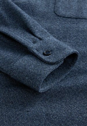 Manșeta cămășii cu model herringbone de culoare bleu închis, cu o textură fină, un închizător cu un singur buton și detalii vizibile de cusătură pe material.