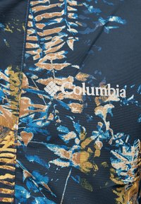 Λογότυπο Columbia σε σκούρο ύφασμα με πολύχρωμο μοτίβο φύλλων και φτέρης σε αποχρώσεις του μπλε, καφέ, κίτρινου και μπεζ.