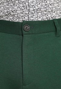 Gröna byxor i strukturerad tyg med knappstängning, detaljering vid midjan och en slät framsida. En blommig skjorta syns ovanför.