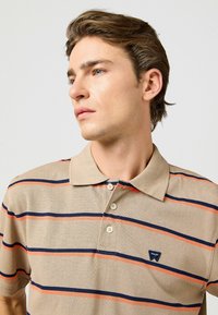 Beige polotröja med horisontella marinblå och orange ränder. Har en klassisk krage och tre knappar vid halsen. Wrangler-logotyp på bröstet.