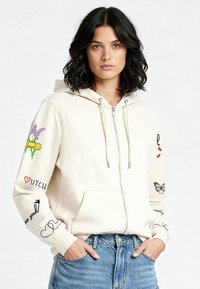 Krema bojeni hoodie s patentnim zatvaračem i džepom tipa kengur, ukrašen šarenim grafičkim detaljima na oba rukava, uključujući cvjetne motive i tekstualne naglaske.